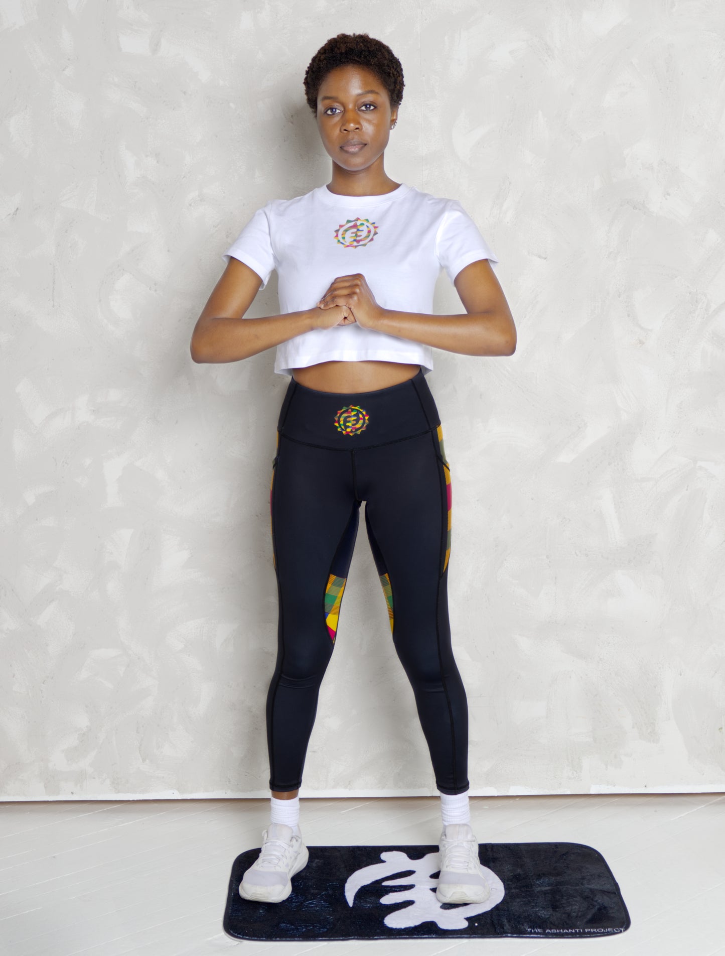 Person wearing a white crop top and black leggings with colorful accents on a light gray background
Black woman wearing white leggings with colorful kente designs on a plain background. Athletic wear. Gym outfit,