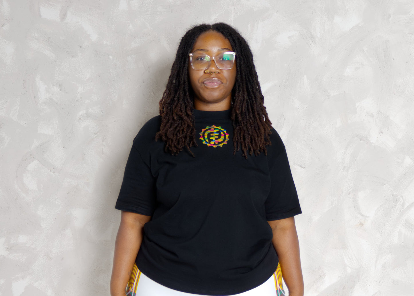 Person wearing a black t-shirt and white leggings with colorful kente patterns, standing on a mat against a light background.