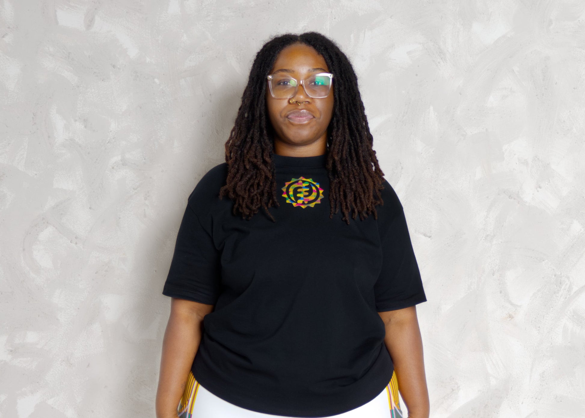 Person wearing a black t-shirt and white leggings with colorful kente patterns, standing on a mat against a light background.
