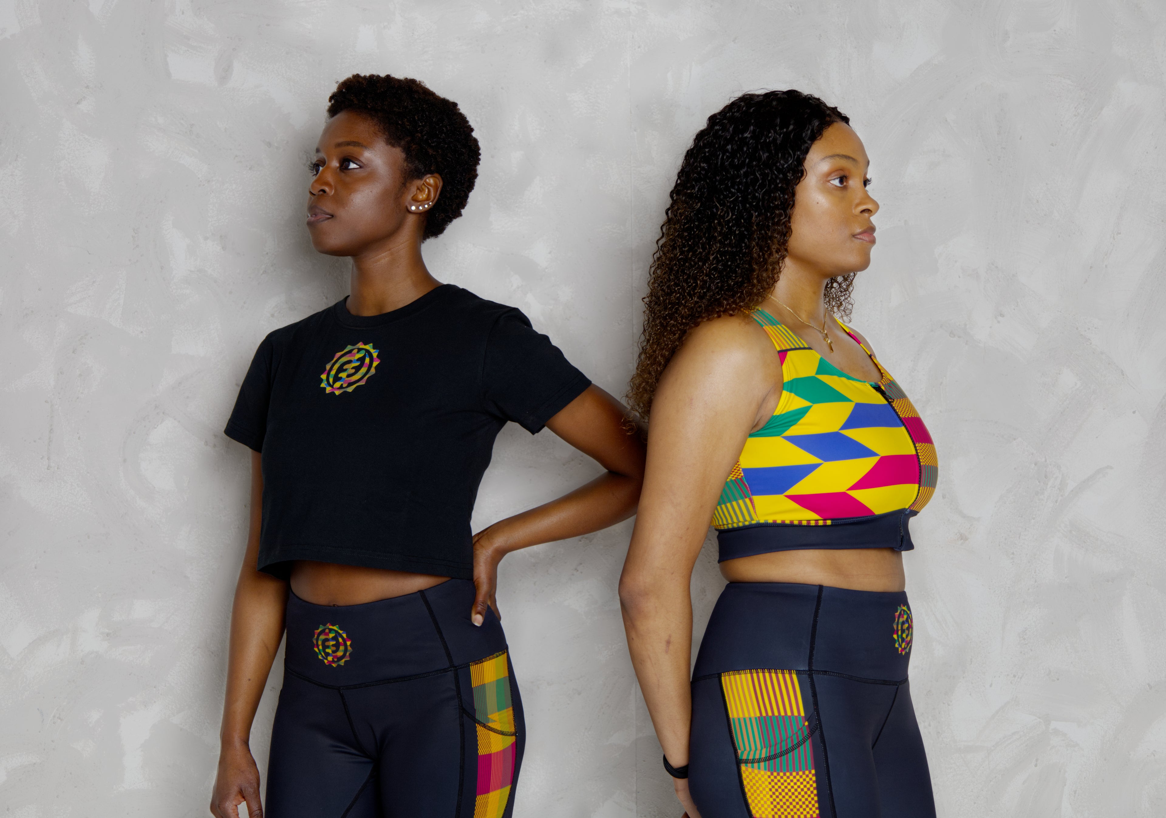 Two women wearing matching outfits with a kente pattern on a plain background