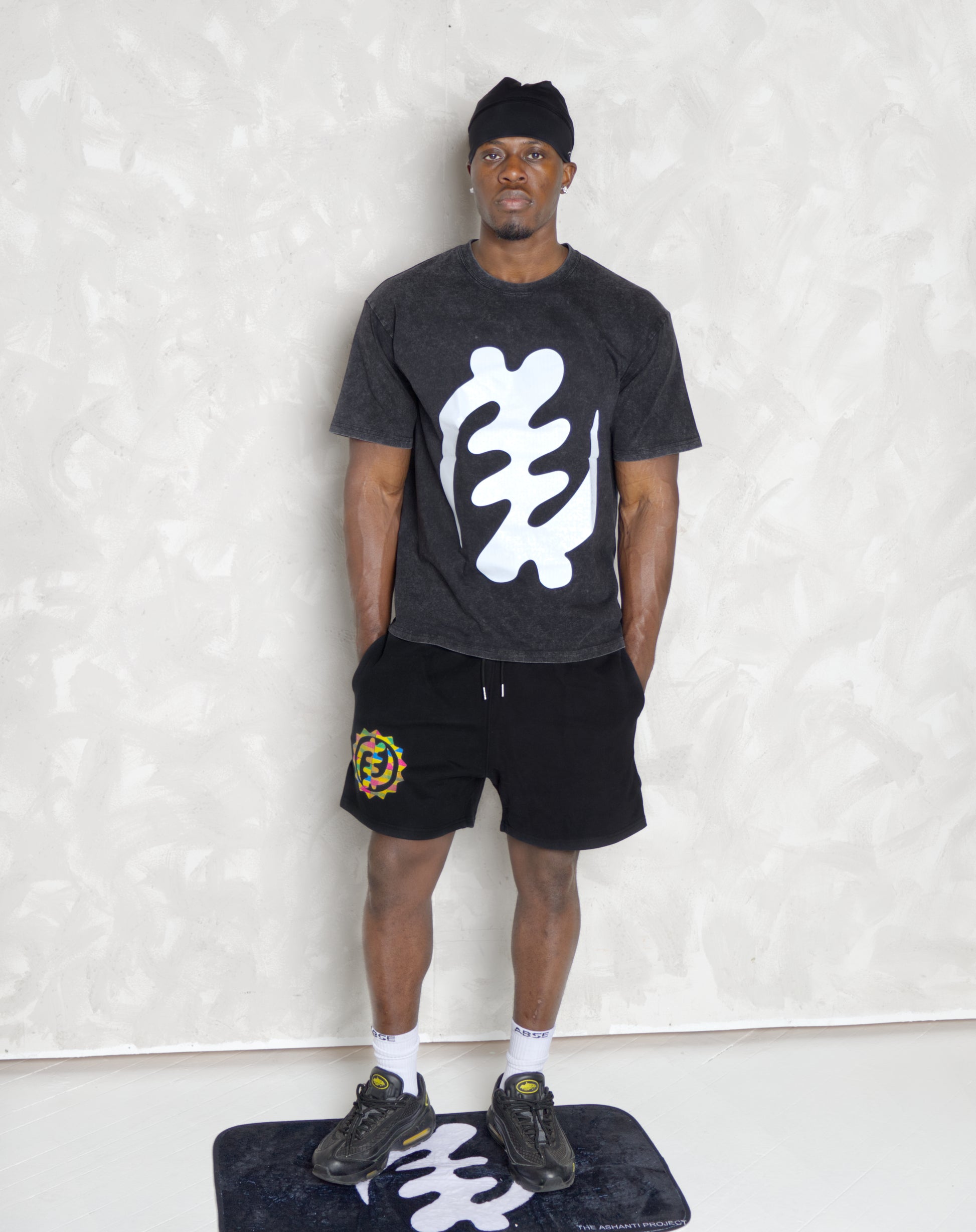 Person wearing a dark t-shirt with a white graphic design, black shorts with a colorful logo, and black sneakers on a light background.