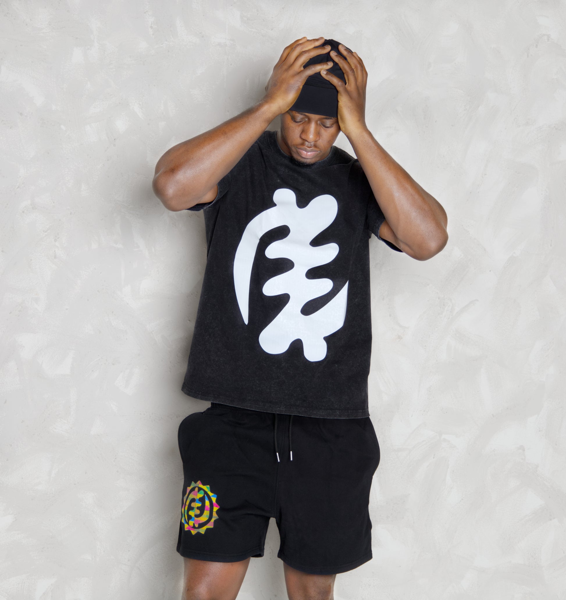 Person wearing a black sleeveless shirt with a white gye nyame design and black shorts with a colorful logo on a light gray background