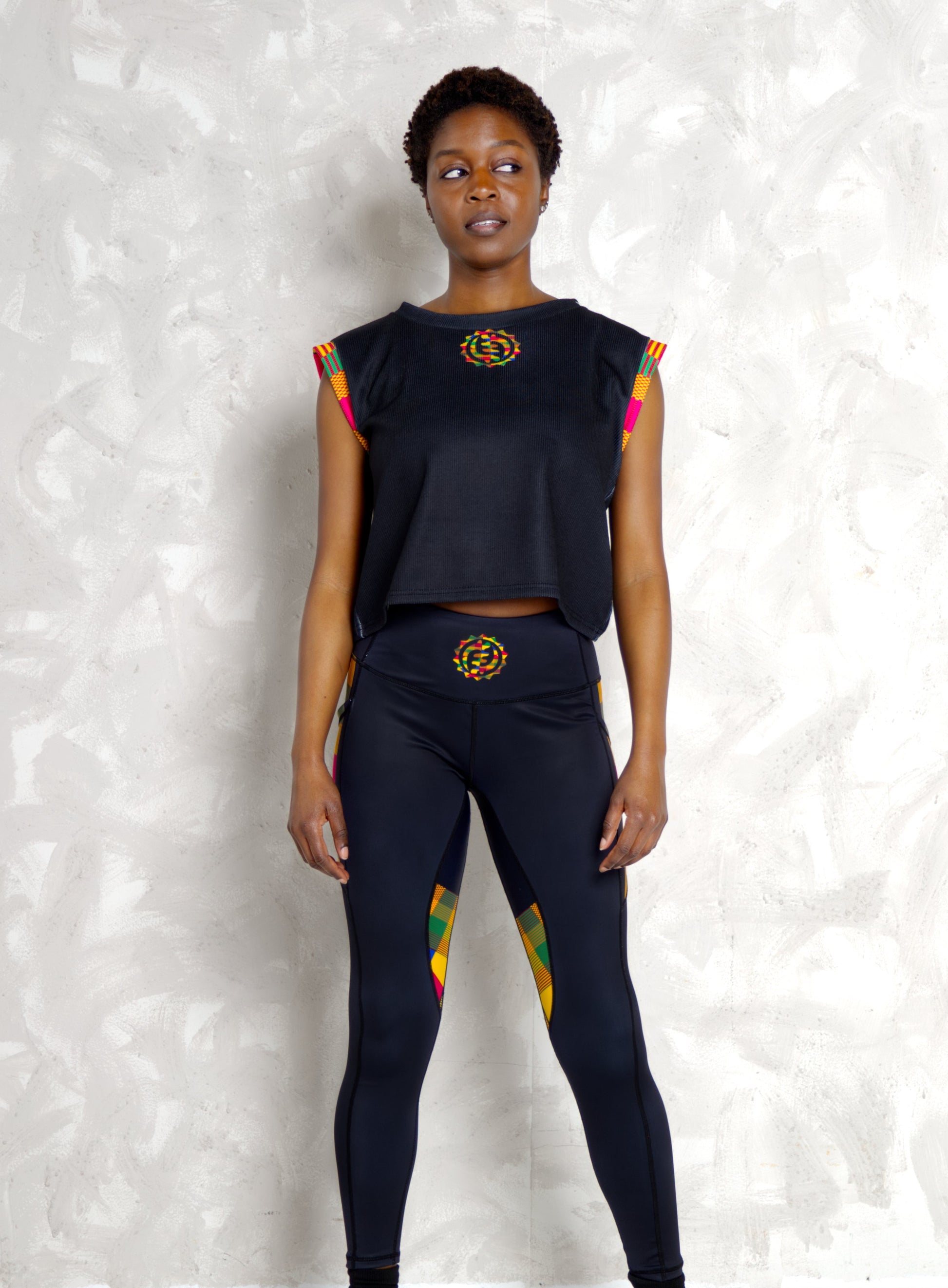 Person wearing a black outfit with colorful Kente accents against a textured white wall.