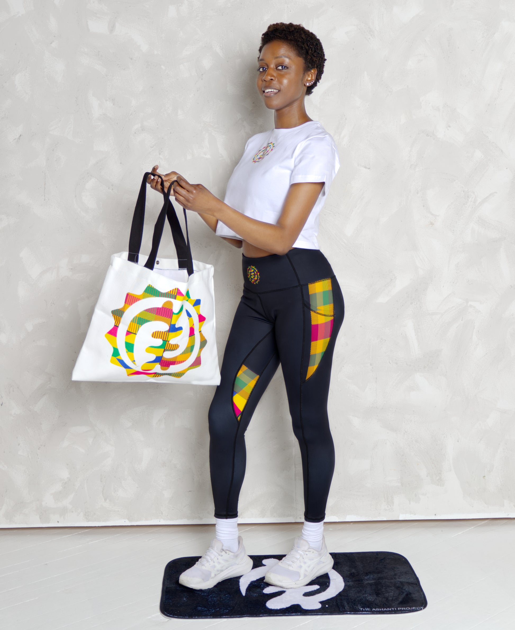 Woman holding a tote bag with a colorful design, wearing black leggings with colorful patterns, on a white background.