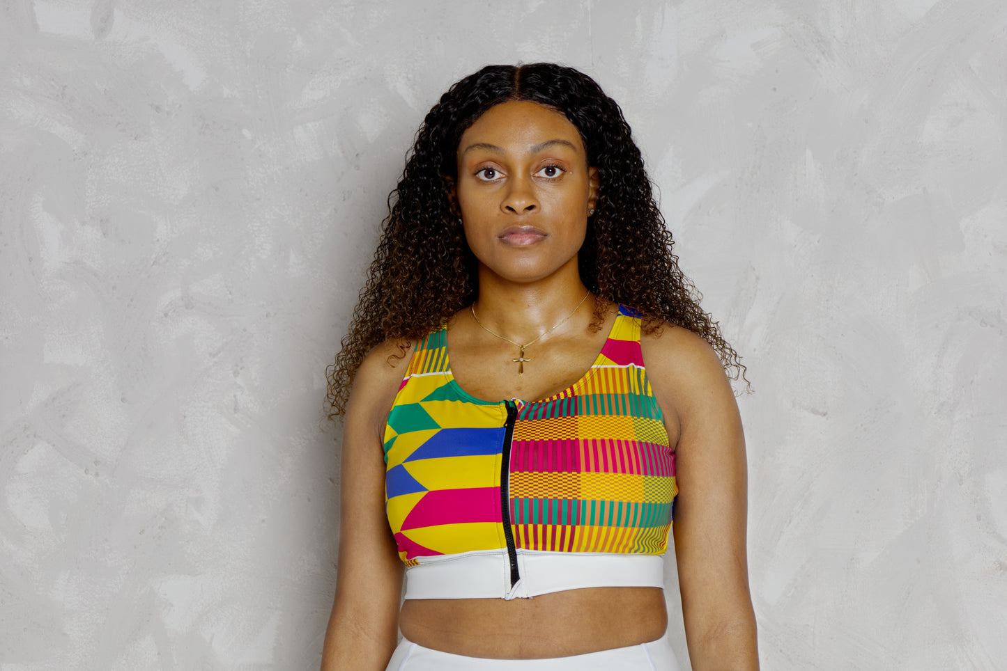 Woman wearing a colorful kente patterned top against a plain background. It’s a padded sports bra with kente designs 