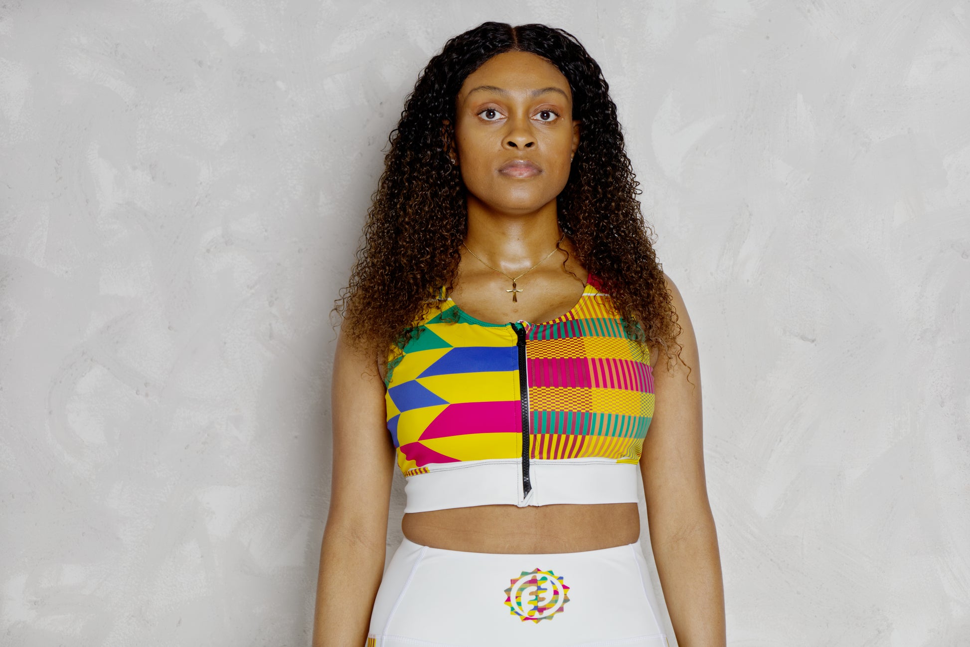 Woman wearing a colorful striped top against a light gray background
Woman wearing a colorful kente patterned top against a plain background. It’s a padded sports bra with kente designs 