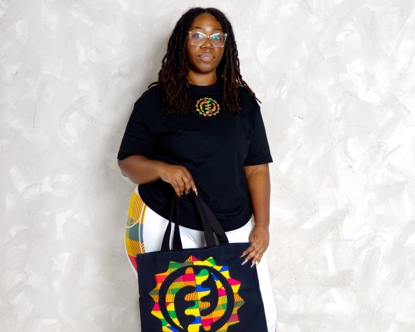 black woman wearing kente leggings and wearing essetial t shirt and holding a kente canvas tote bag
