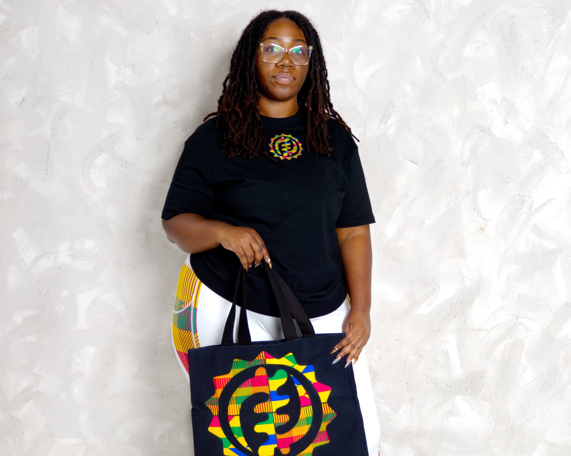black woman wearing kente leggings and wearing essetial t shirt and holding a kente canvas tote bag